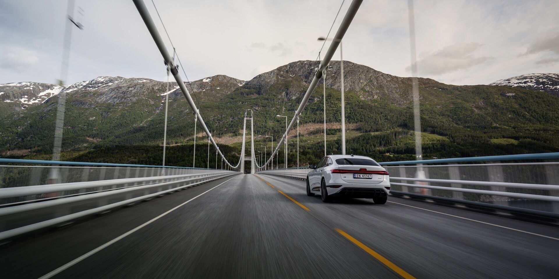 All charged up: from Copenhagen to Bergen in the A6 e-tron - An Audi A6 Sportback e-tron in Glacier white metallic drives over the Hardanger Bridge in Norway. Wooded mountains are visible in the background.