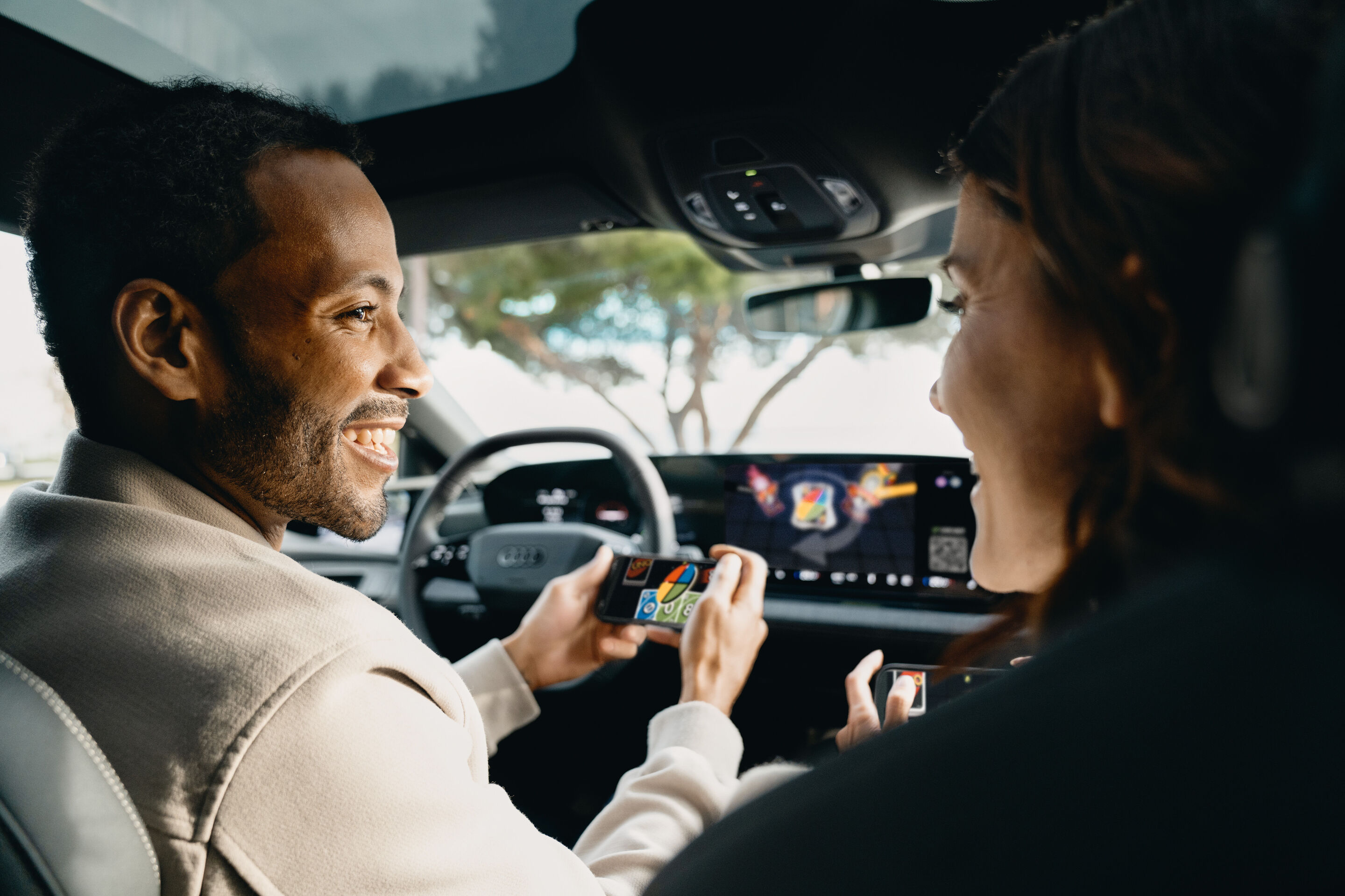 Gaming fun on the go: Audi integrates AirConsole - A game from the AirConsole gaming platform runs on the touch display in an Audi. The driver and passenger smile at each other and each hold a smartphone in their hands. They use the smartphones as controllers...